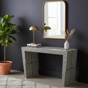 May include: A black and white striped console table with a gold-toned lamp, mirror, and a small white vase with dried plants. The table has a geometric pattern and is placed against a dark gray wall.