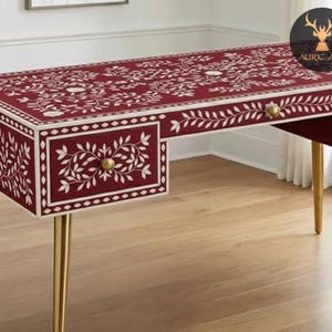 Puede incluir: Un escritorio burdeos con incrustaciones florales blancas y patas doradas. El escritorio tiene dos cajones con pomos dorados y un patrón floral decorativo en la parte superior y los lados. Un mueble elegante.