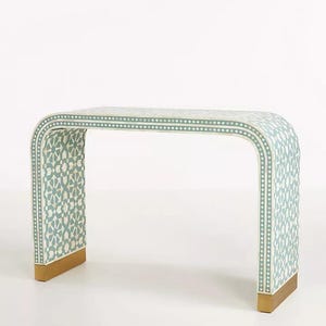 May include: A decorative, curved console table with a light blue and white geometric pattern. The table has a gold-coloured base and measures approximately 122 cm long, 41 cm wide, and 76 cm high.