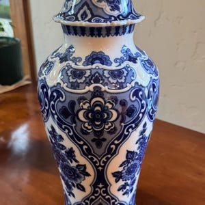 May include: A blue and white ceramic vase with a lid. The vase features a floral pattern in shades of blue against a white background. The design includes flowers and decorative scrollwork. The vase is a decorative home decor item.