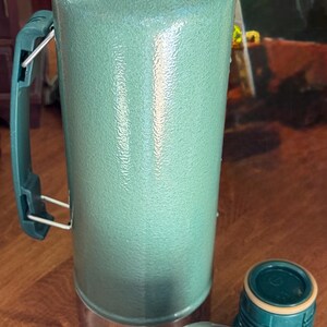 May include: A green insulated thermos with a stainless steel lid and handle. Two additional stainless steel and green caps are next to the thermos. The thermos is designed to keep liquids hot or cold for extended periods.