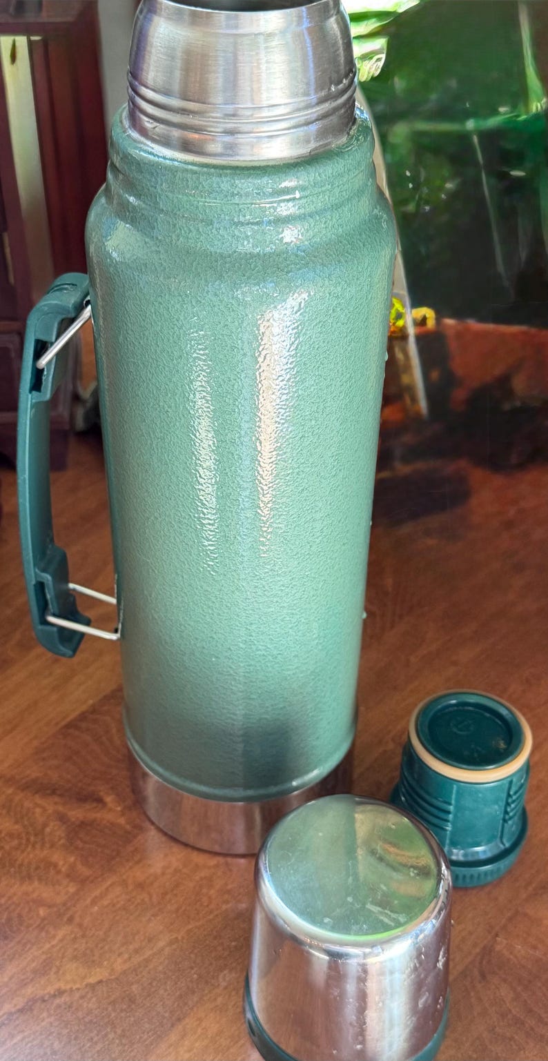 May include: A green insulated thermos with a stainless steel lid and handle. Two additional stainless steel and green caps are next to the thermos. The thermos is designed to keep liquids hot or cold for extended periods.