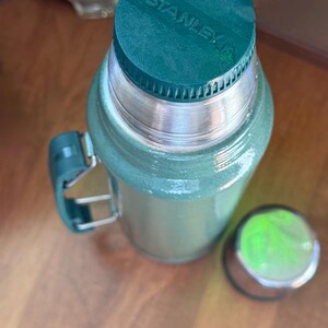 May include: Green Stanley thermos with a silver rim and handle. The thermos has a matching green lid with the brand name "STANLEY" printed on it. The lid is off to the side. The thermos is on a wooden surface.