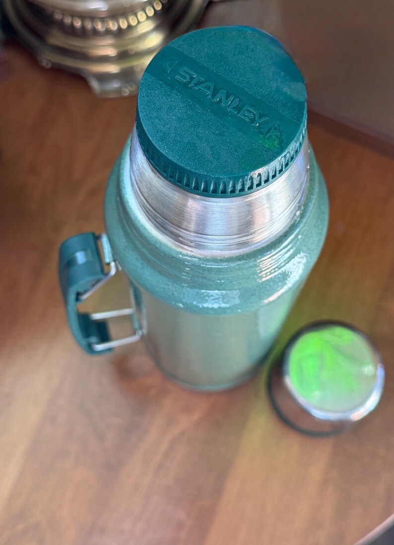 May include: Green Stanley thermos with a silver rim and handle. The thermos has a matching green lid with the brand name "STANLEY" printed on it. The lid is off to the side. The thermos is on a wooden surface.