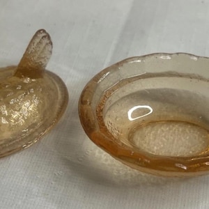 May include: A small, amber-colored glass dish and a lid with a handle. The dish has a scalloped edge. The lid has a textured surface and a handle that resembles a bird's head.