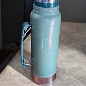 May include: A teal-colored thermos with a silver lid and a copper-colored base. The thermos has a blue handle and a green band near the top. The thermos is designed to keep liquids hot or cold.