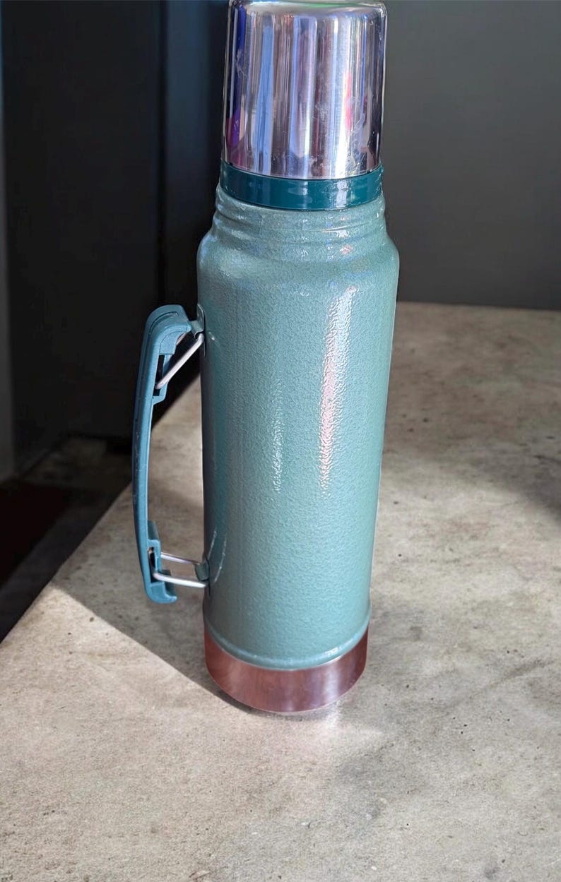 May include: A teal-colored thermos with a silver lid and a copper-colored base. The thermos has a blue handle and a green band near the top. The thermos is designed to keep liquids hot or cold.