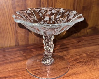 May include: Clear glass pedestal bowl with a scalloped rim and etched floral design. The bowl sits on a decorative stem and a round base. The bowl is likely used for serving desserts or displaying decorative items.
