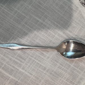 Op de afbeelding: Een zilveren lepel met een druppelvormige kom en een lange, slanke steel. De lepel ligt op een wit, getextureerd oppervlak met een kanten rand.