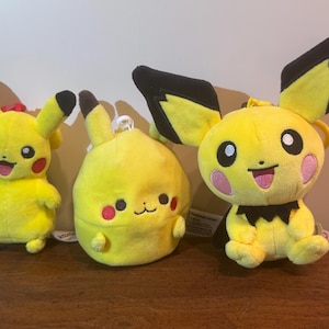 May include: Three yellow Pikachu plush toys with black-tipped ears, red cheeks, and smiling faces. One has a red loop, another is round, and the third has large ears. They are sitting on a brown surface.