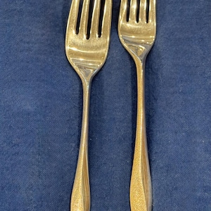 May include: Two gold-coloured forks with a textured finish. The forks have four tines and a slender, diamond-shaped handle. The forks are displayed against a dark blue background.