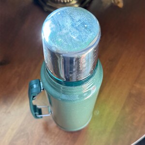 May include: A vintage green thermos with a silver lid and a green handle. The thermos is cylindrical and appears to be made of metal. The handle is attached to the side of the thermos. The thermos is sitting on a wooden surface.