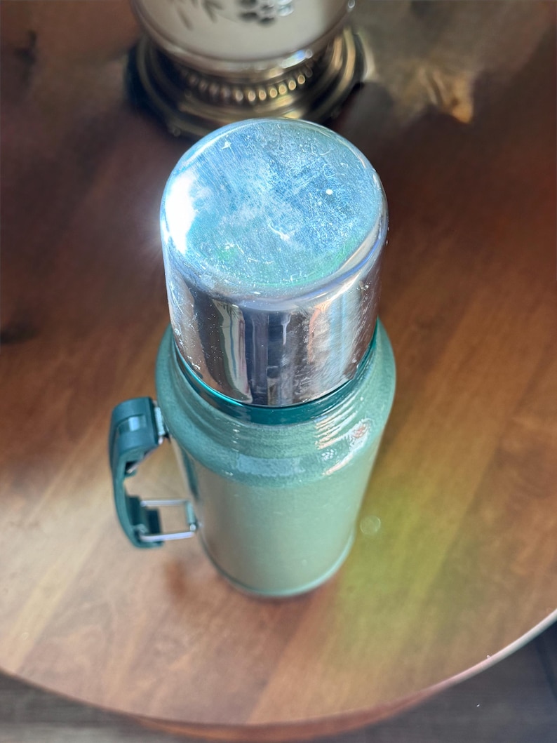 May include: A vintage green thermos with a silver lid and a green handle. The thermos is cylindrical and appears to be made of metal. The handle is attached to the side of the thermos. The thermos is sitting on a wooden surface.