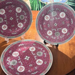 May include: Three antique decorative plates with a deep red background and intricate floral and geometric patterns. Each plate features white and red Chinese characters and a turquoise and white border. The plates are displayed on clear stands.