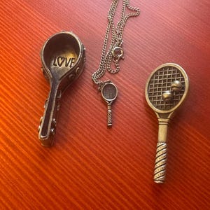 May include: Three bronze-toned tennis-themed jewelry pieces on a reddish-brown surface. Includes a spoon-shaped charm with "LOVE" inscribed, a tennis racket pendant on a chain, and a tennis racket charm with two balls.