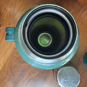 May include: An overhead view of a green Stanley thermos with a silver rim and a small handle. The interior is black and green. A small, silver lid sits next to the thermos.