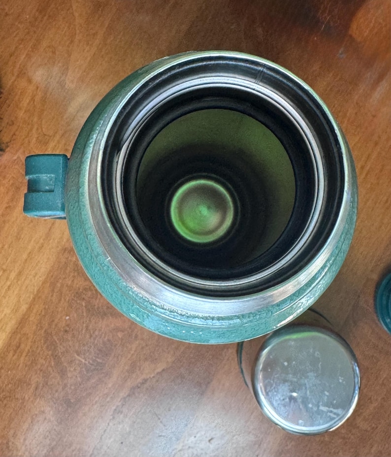 May include: An overhead view of a green Stanley thermos with a silver rim and a small handle. The interior is black and green. A small, silver lid sits next to the thermos.