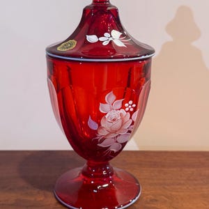 May include: A ruby red glass jar with a lid, featuring a white floral design. The jar has a pedestal base and a decorative finial on top. The floral design includes a rose and other delicate flowers. The jar is likely used for storage or display.