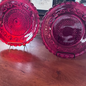 Puede incluir: Dos platos de cristal rojo rubí con diseños ornamentados en relieve y bordes festoneados. Los platos se exhiben sobre soportes de metal negro, mostrando sus intrincados patrones. Los platos son probablemente para fines decorativos o de servicio.
