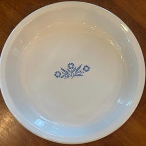 May include: White ceramic pie plate with a blue floral design in the center. The plate has a wide rim and a slightly curved shape. The floral design features small blue flowers and leaves.