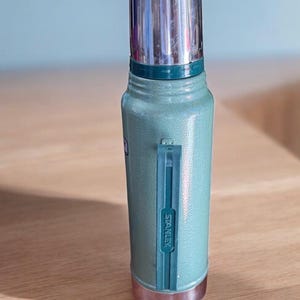 May include: A light green Stanley thermos with a silver lid and base. The thermos has a handle and the brand name "Stanley" is printed on the side. The thermos is made of metal and is designed to keep liquids hot or cold.