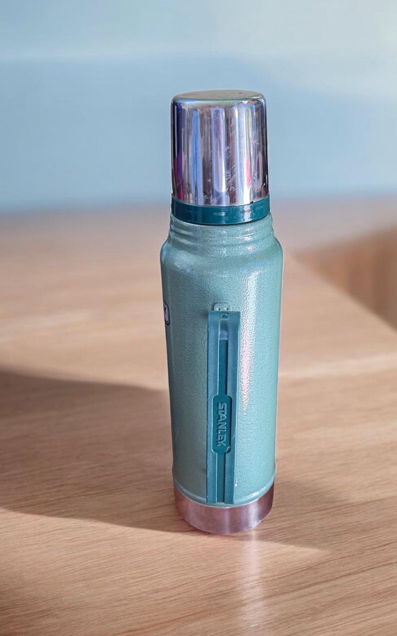 May include: A light green Stanley thermos with a silver lid and base. The thermos has a handle and the brand name "Stanley" is printed on the side. The thermos is made of metal and is designed to keep liquids hot or cold.