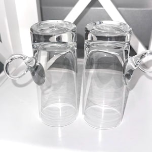 Könnte beinhalten: Zwei transparente Glasbecher mit abgerundeten Griffen. Die Becher sind zylindrisch mit leicht ausgestelltem Rand und dickem, flachem Boden. Die Griffe sind an der Seite des Bechers befestigt. Die Becher stehen auf einer weißen Oberfläche.