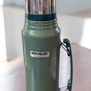 May include: A green Stanley thermos with a silver lid and handle. The thermos has a textured surface and the Stanley logo is visible. The thermos is on a wooden surface.