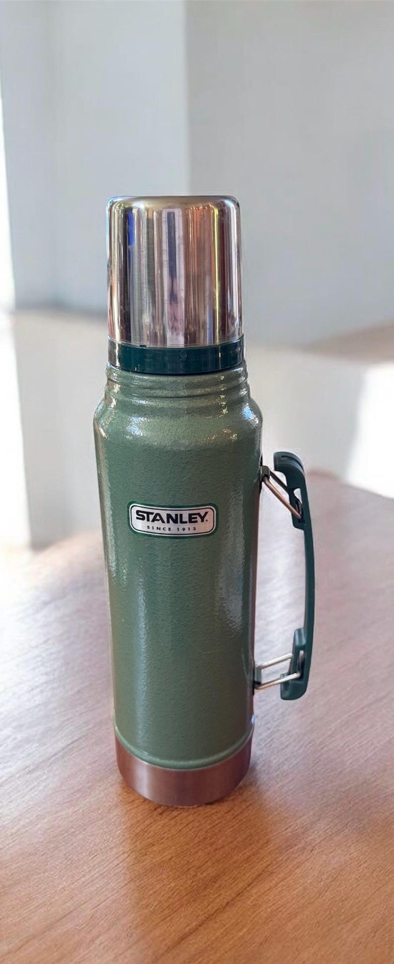 May include: A green Stanley thermos with a silver lid and handle. The thermos has a textured surface and the Stanley logo is visible. The thermos is on a wooden surface.