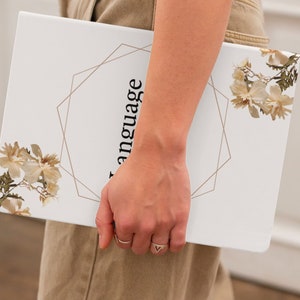 May include: A white notebook with a gold geometric design and the word "language" written vertically on the cover. The notebook is decorated with dried flowers.