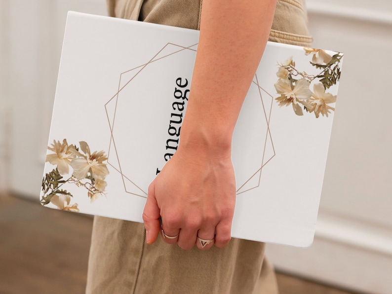 May include: A white notebook with a gold geometric design and the word "language" written vertically on the cover. The notebook is decorated with dried flowers.