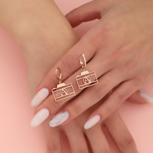 Puede incluir: Pendientes colgantes en forma de cámara de oro rosa. Los pendientes están en la mano de una modelo con esmalte de uñas blanco.