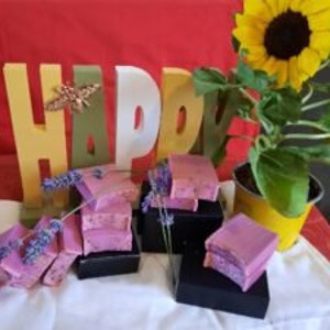 May include: A stack of handmade lavender soap bars in a purple and white swirl pattern, displayed on a white cloth with a wooden sign that says "Happy" and a sunflower in a yellow pot.
