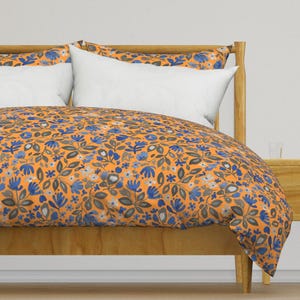 Puede incluir: Una cama con una funda nórdica y fundas de almohada con estampado floral naranja. La ropa de cama presenta un patrón repetido de flores y hojas azules, grises y blancas. La estructura de la cama es de madera clara.