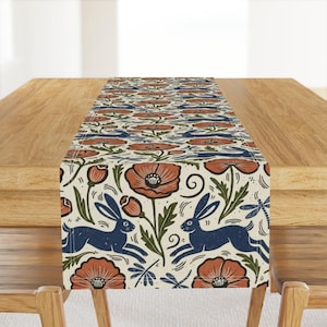 Può includere: Runner da tavolo color crema con un motivo ripetuto di papaveri arancioni, conigli blu e foglie verdi. Il runner è posto su un tavolo di legno, mostrando il suo design decorativo. Il runner è largo circa 30 cm.
