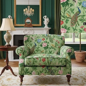 Op de afbeelding: Een groene fauteuil met een bloemen- en pauwenpatroon. De stoel heeft een afgeronde rug en armleuningen, met donkere houten poten. De achtergrond omvat een tafellamp, een bijzettafel en een open haard.