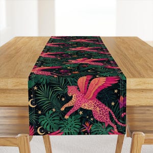 Puede incluir: Camino de mesa decorativo con un diseño de leopardo alado rosa sobre un fondo de selva tropical. El camino de mesa tiene una base verde oscuro con detalles en rosa, naranja y blanco. El diseño incluye estrellas y lunas.