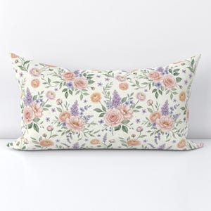 Puede incluir: Un cojín rectangular con un patrón repetitivo de acuarela que muestra rosas de color rosa suave, ranúnculos naranjas y ramas de lilas moradas con hojas verdes sobre un fondo blanco roto. El diseño floral está distribuido por la tela.