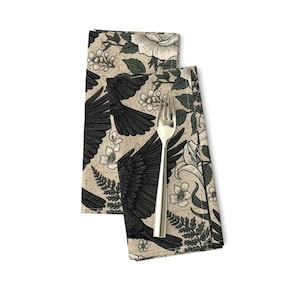 May include: Two beige cloth napkins with a black raven and floral pattern. The napkins feature a repeating design of raven wings, white flowers, and green leaves. A silver fork rests on the napkins.