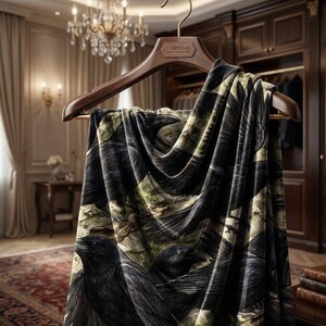 May include: A dark scarf with a raven print hangs from a wooden hanger. The scarf features a repeating pattern of black ravens against a backdrop of muted green and brown tones. The hanger has the text "CORVIDAE" on it.