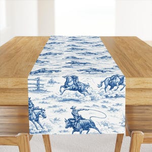 May include: A blue and white table runner with a Western-themed toile pattern. The design features cowboys on horseback, roping cattle, and other ranch scenes. The runner is placed on a wooden table.
