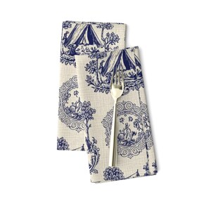 May include: Two folded cream-colored cloth napkins with a blue toile pattern featuring tents, trees, and figures. A silver fork rests on top of one napkin. The napkins are likely for a dining table setting.