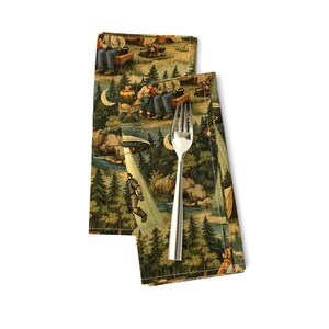 May include: Two folded cloth napkins with a whimsical forest scene featuring a campfire, people, a UFO, and a figure in a spacesuit. A silver fork rests on top of the napkins. The napkins have a green, brown, and beige color scheme.
