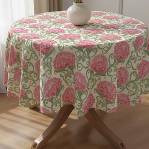 Könnte beinhalten: Ein runder Tischläufer mit einem floralen Muster aus rosa Hortensien und grünen Blättern auf cremefarbenem Hintergrund. Der Tischläufer liegt auf einem Holztisch, mit einer weißen Vase darauf. Das Design ist ein klassisches Blumenmuster.