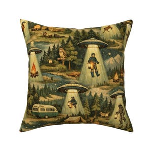 May include: Cushion with a quirky forest scene featuring UFOs, campers, and wildlife. The pillow has a tapestry-like design with a green, brown, and blue colour palette. The design includes a camper van, a tent, and a campfire.