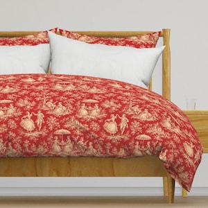May include: A red and cream duvet cover featuring a repeating toile de Jouy pattern of figures in 18th-century attire, including people dancing and sitting under umbrellas, set against a wooden bed frame with two plain white rectangular pillows.
