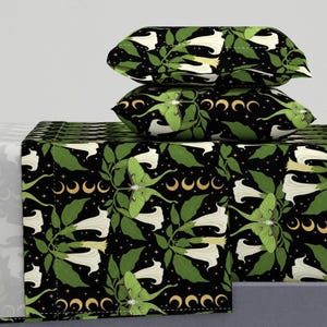 Puede incluir: Juego de cama negro con un patrón repetido de flores blancas en forma de trompeta, polillas verdes y lunas crecientes doradas. El juego incluye dos fundas de almohada y una sábana bajera. El fondo es negro con pequeñas estrellas blancas.