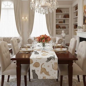 Könnte beinhalten: Ein Esstisch, der für eine Mahlzeit gedeckt ist, mit einem weißen Tischläufer mit Katzenillustrationen in verschiedenen Posen. Der Tisch ist von gepolsterten Stühlen umgeben und mit Tellern und Gläsern gedeckt. Ein Kronleuchter hängt darüber, und ein Blumenarrangement befindet sich in der Mitte.