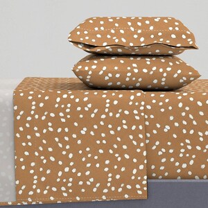 Puede incluir: Un juego de cama con un estampado de lunares marrones y blancos. El juego incluye dos fundas de almohada y una sábana bajera. Las fundas de almohada están apiladas encima de la sábana bajera. La combinación de colores es marrón cálido con lunares blancos.