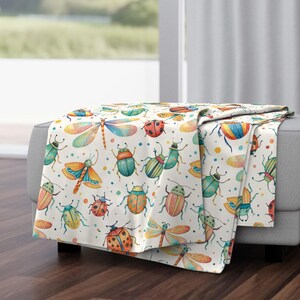 Op de afbeelding: Een witte deken met een kleurrijk aquarelpatroon van diverse insecten, waaronder libellen, lieveheersbeestjes en kevers. De insecten zijn in tinten blauw, groen, oranje en geel, met kleine gekleurde stippen.